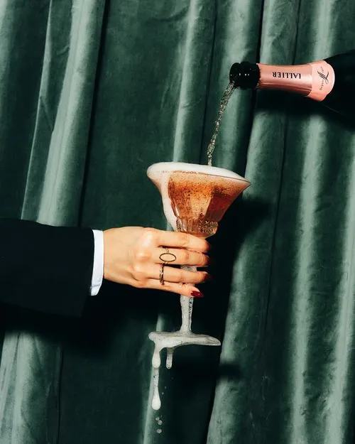 Hand with rings and red nail polish holding a champagne glass as sparkling wine is poured, causing foam to overflow.