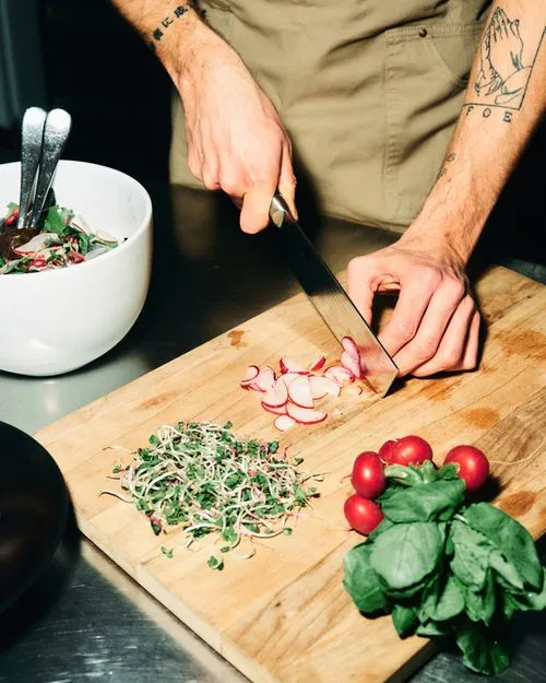 Person chopping sliced radishes on a wooden cutting board with microgreens and cherry tomatoes nearby.