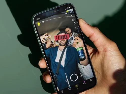 Hand holding a smartphone displaying a Snapchat photo of a man holding a beer bottle at a party with a '3 LINDEN' label over his eyes.