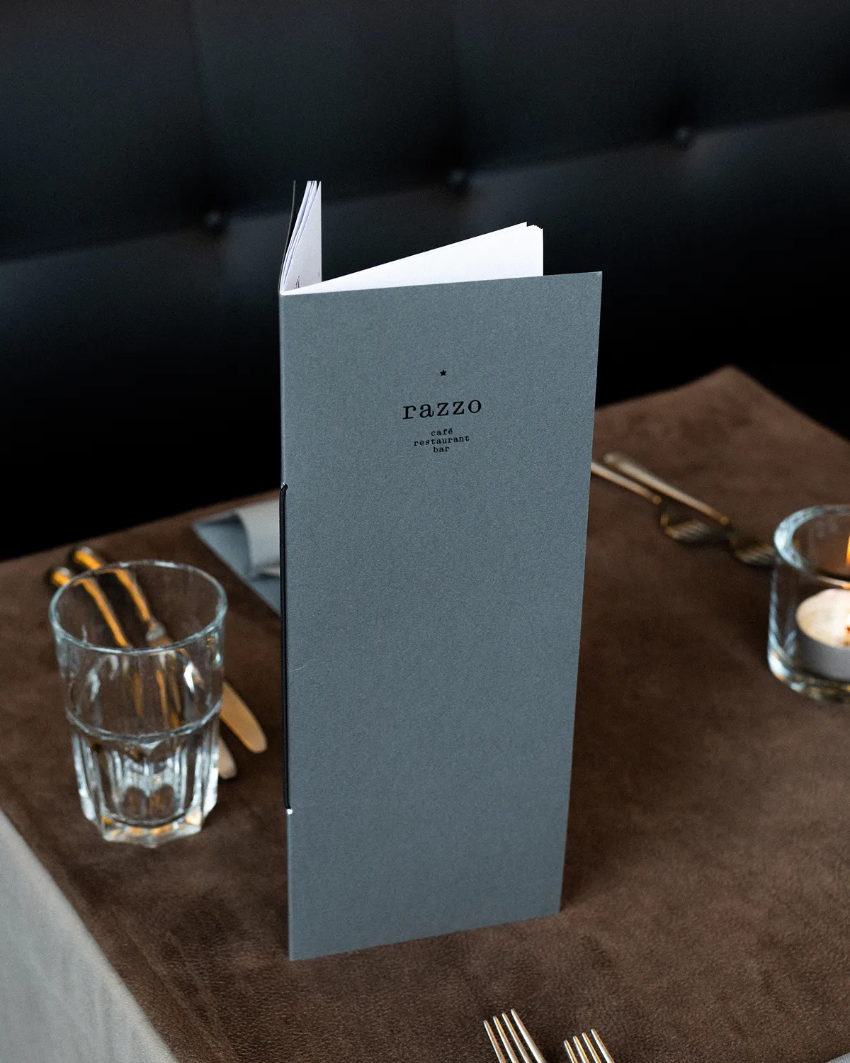 Tall gray menu card with 'razzo ace restaurant bar' text on a brown tablecloth with utensils, glass, and candle in the background.