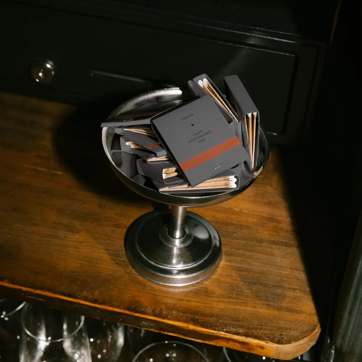 Metal pedestal bowl filled with black matchboxes with a brown stripe, placed on a wooden surface.