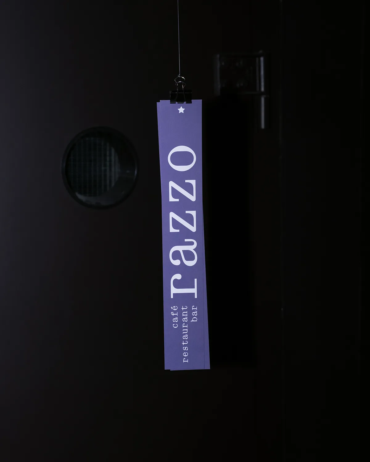 Purple rectangular sign hanging by a clip with the text 'café restaurant bar razzo' on a dark background.