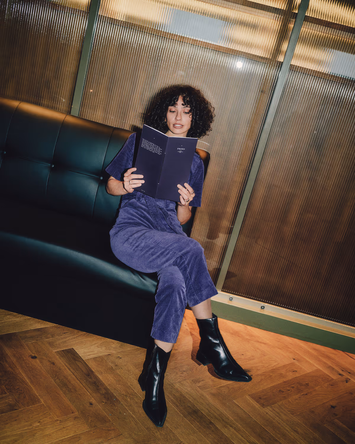 Person with curly hair sitting on a dark green leather couch, reading a dark blue menu, wearing a matching purple corduroy outfit and black ankle boots.