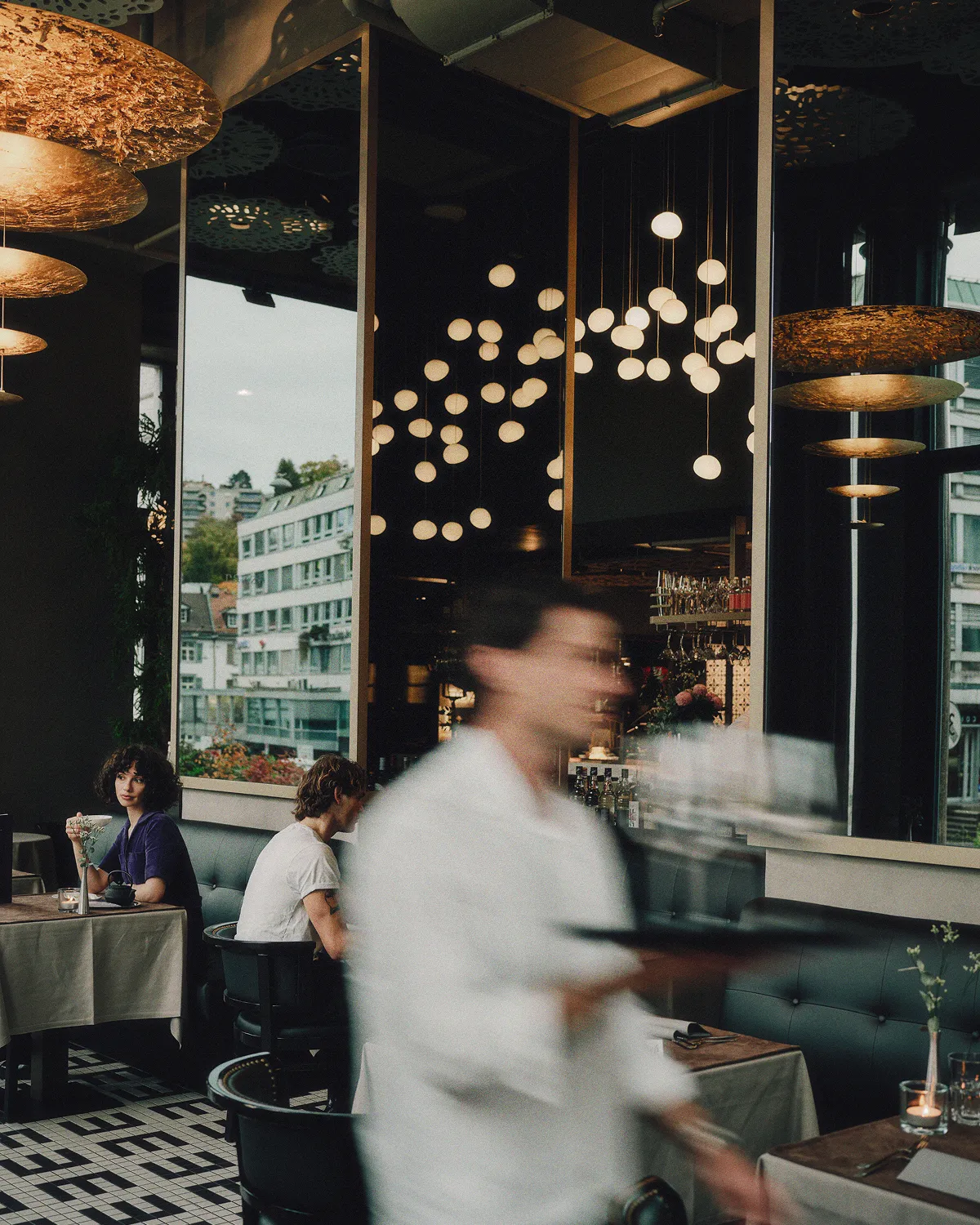 Blurry waiter carrying drinks past two patrons seated at tables in a stylish restaurant with modern pendant lights and large windows showing urban buildings outside.