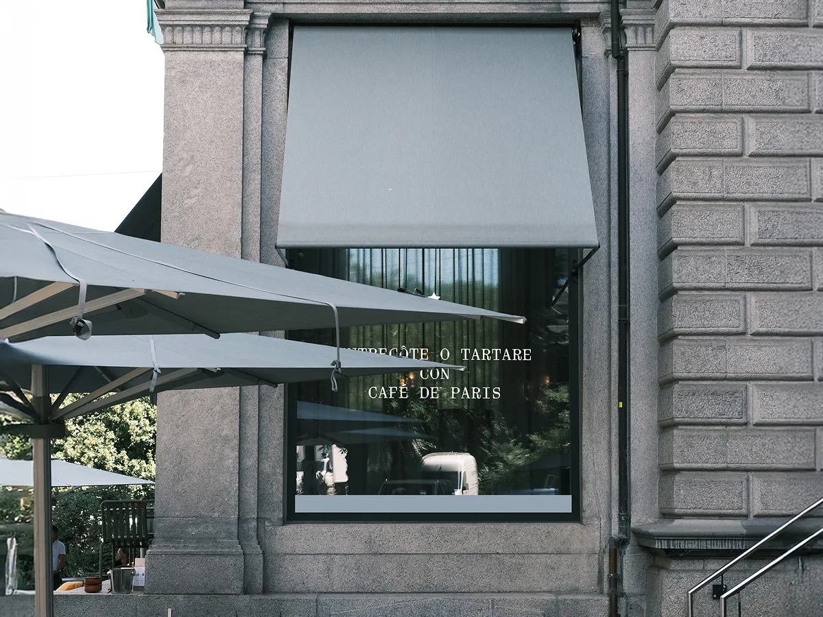 Exterior of a stone building with a large window covered by a gray awning, featuring text about a tartare recipe with café de Paris sauce, and outdoor umbrellas partially visible on the left.