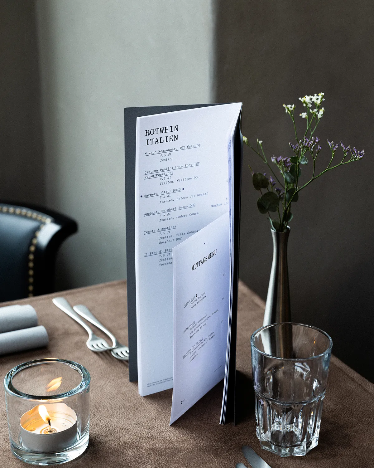 Table set with a lit candle in a glass holder, a folded menu labeled 'ROTWEIN ITALIEN', a clear glass tumbler, silver cutlery, and a tall vase with small white and purple flowers.