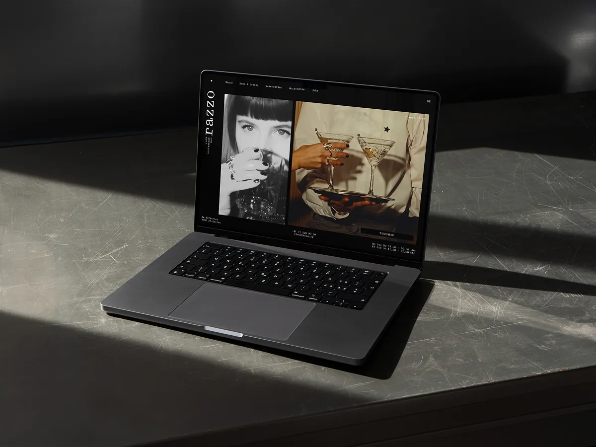 Open laptop on a gray table displaying a website with a black-and-white photo of a woman drinking and a color photo of hands holding martini glasses.