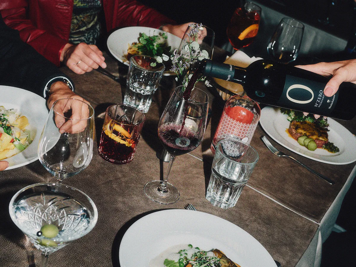 People dining together at a table with plates of food, glasses of water, cocktails, and a bottle of red wine being poured into a wine glass.