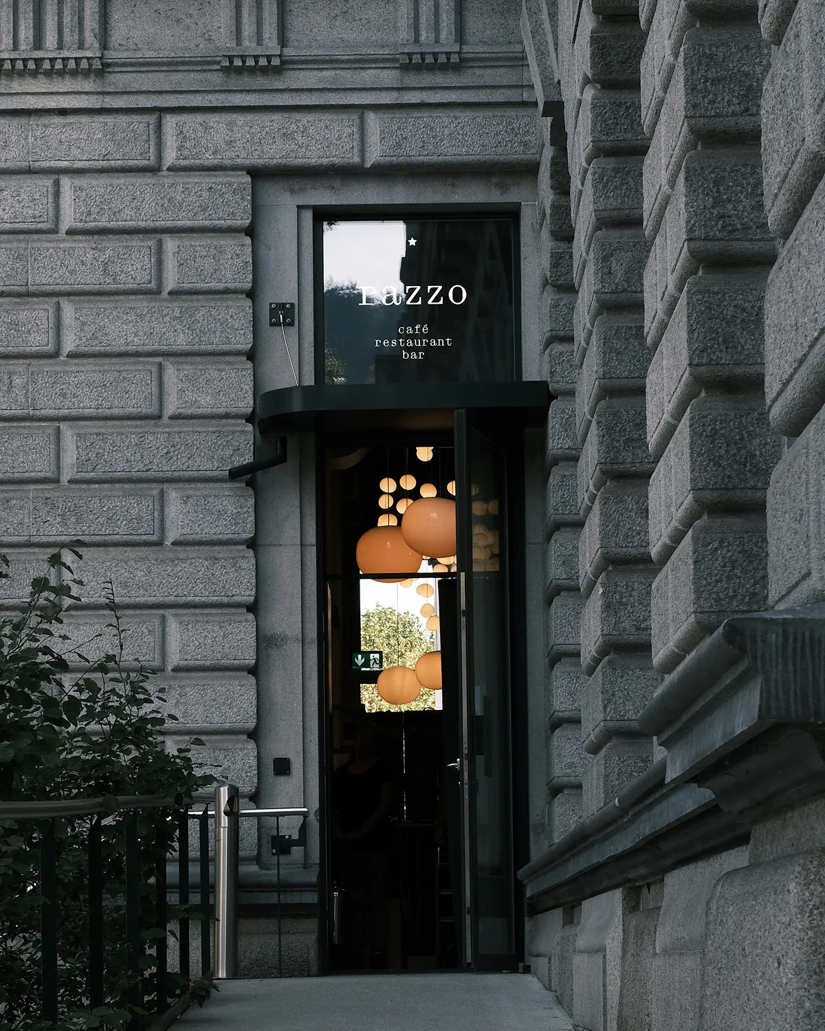 Entrance of a stone building with a glass door showing glowing round pendant lights inside and sign reading 'Lazzo café restaurant bar.'