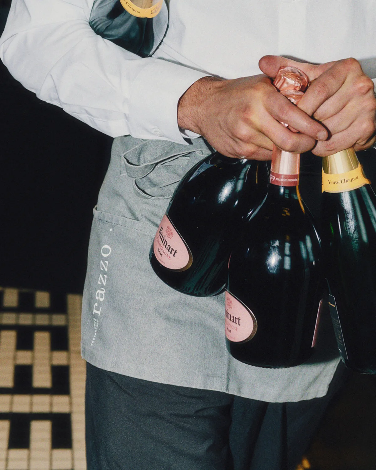 Server wearing a gray Cazzo apron holding three bottles of champagne, two Ruinart and one Veuve Clicquot.