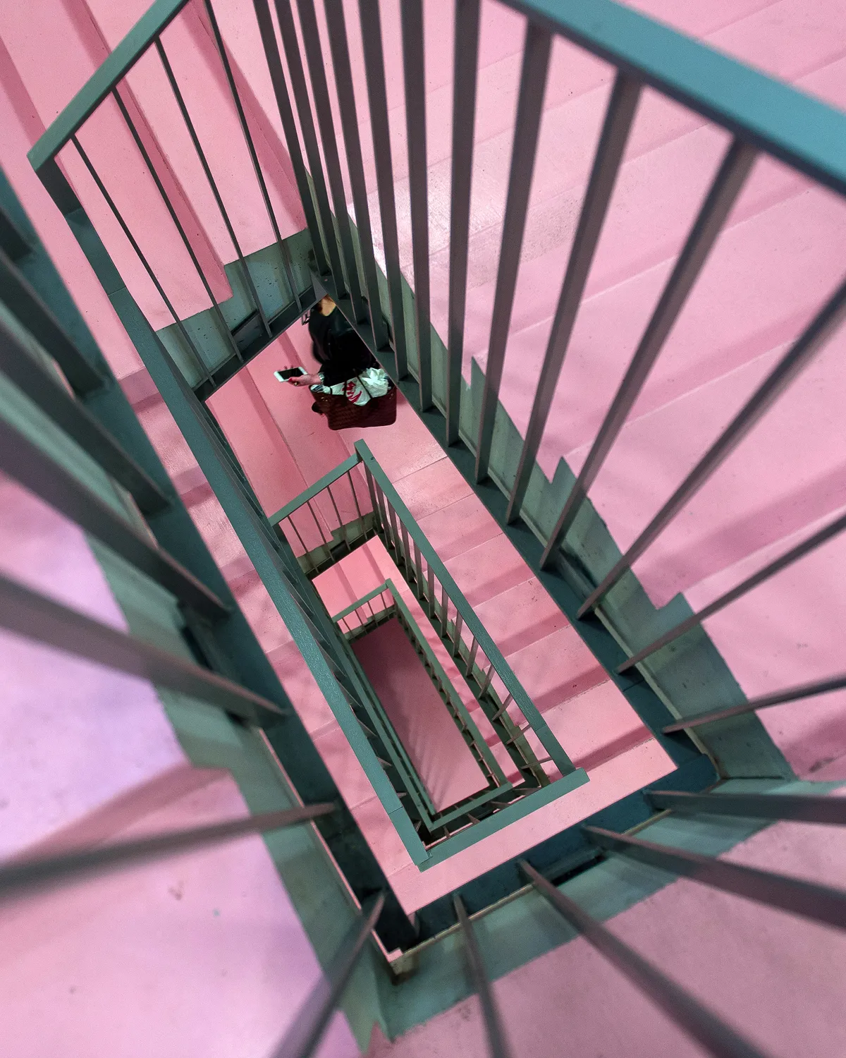 Top-down view of a metal stairwell with pink walls and stairs, featuring a person holding a phone going down.