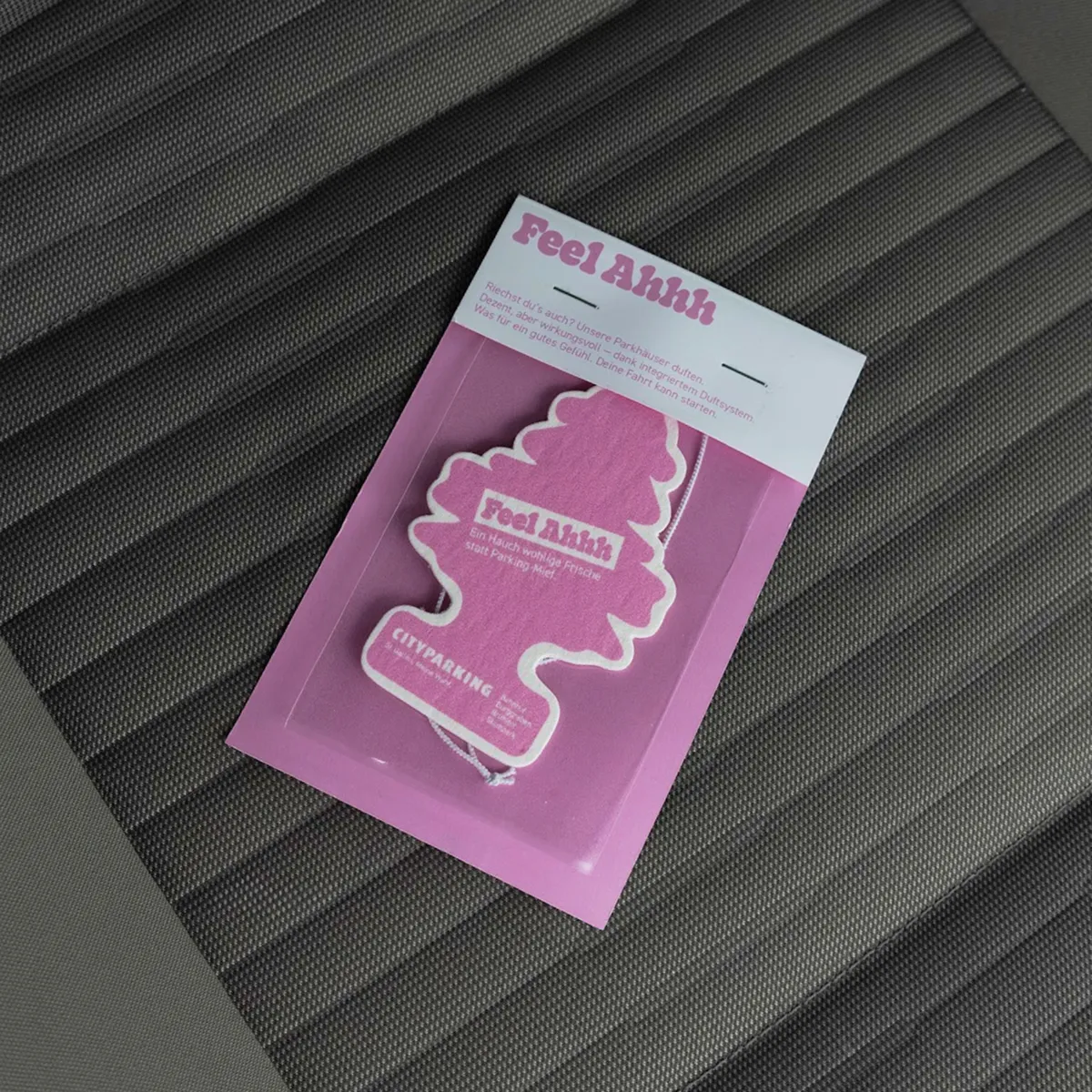 Pink tree-shaped car air freshener labeled 'Feel Ahhh' on a textured dark car seat.