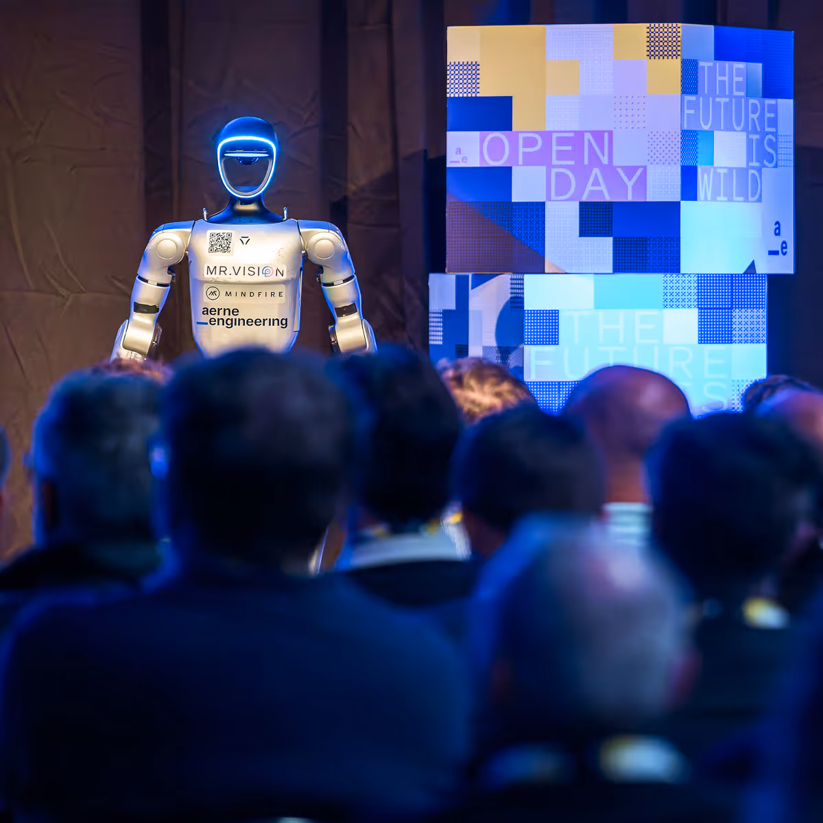 Robot speaker with glowing face presenting to an audience at an event with a futuristic screen backdrop.