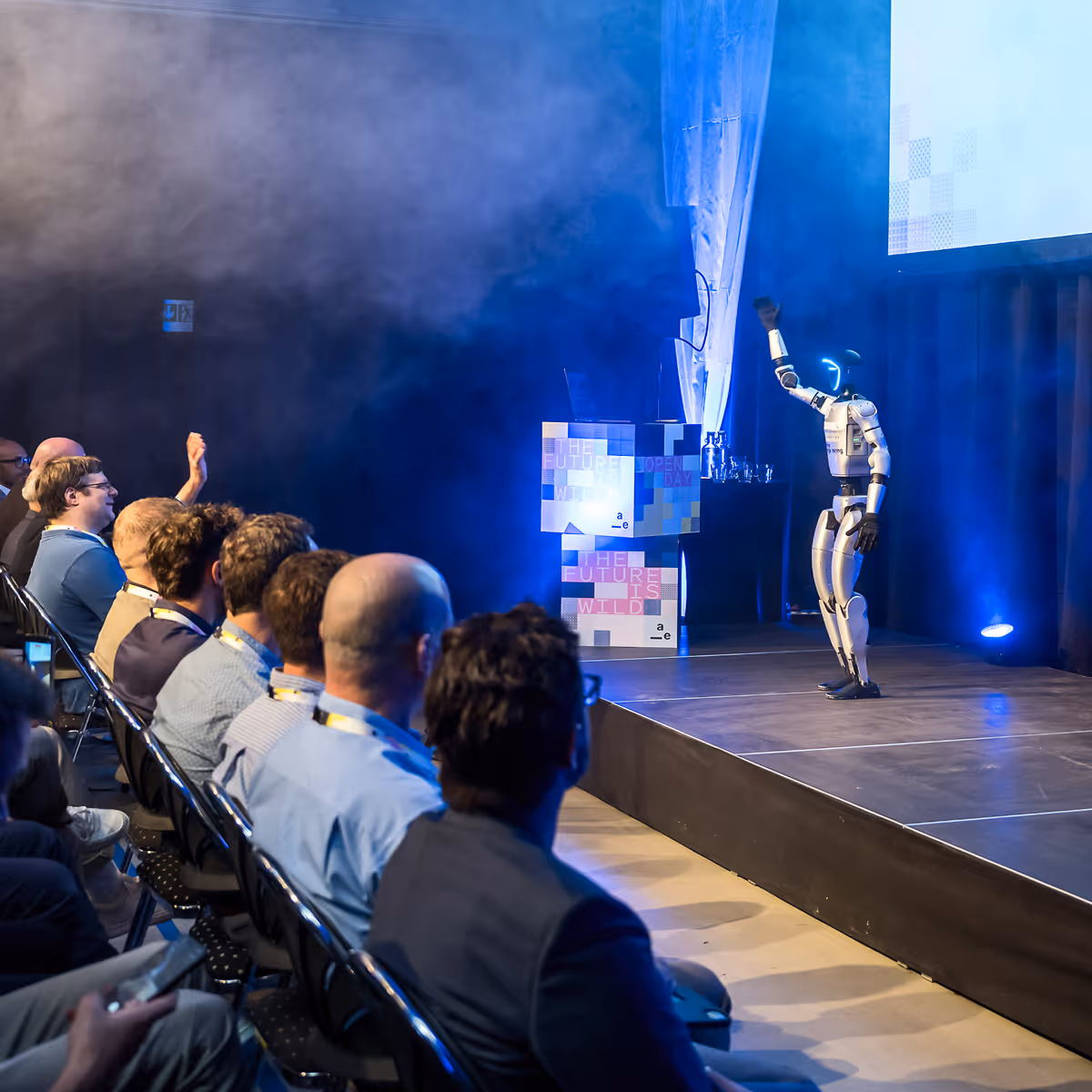 Humanoid robot raises fist on stage while an audience watches and applauds at a technology event.