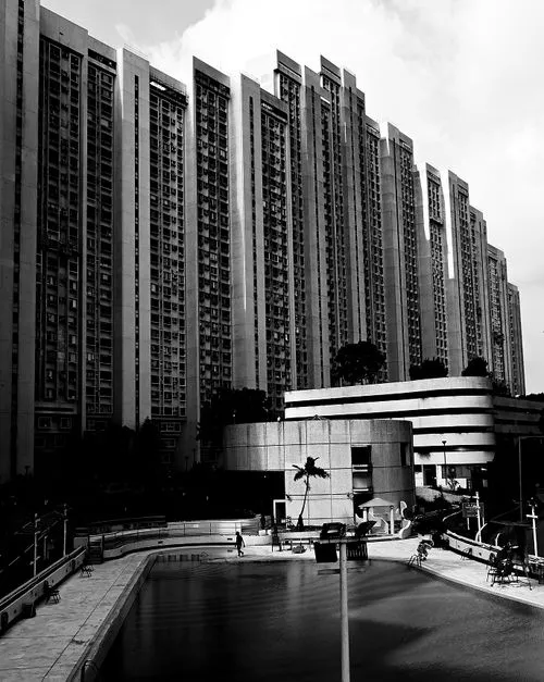 Black and white photo of a large high-rise apartment building with a swimming pool in the foreground and a person walking near the pool.