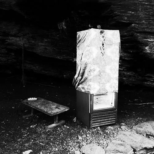 Old refrigerator covered with a worn cloth and a rustic wooden bench nearby on rocky ground inside a dark cave or rock shelter.