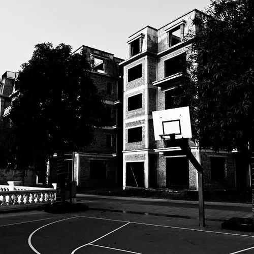 Empty outdoor basketball court with a single hoop in front of an unfinished multi-story brick building and trees.