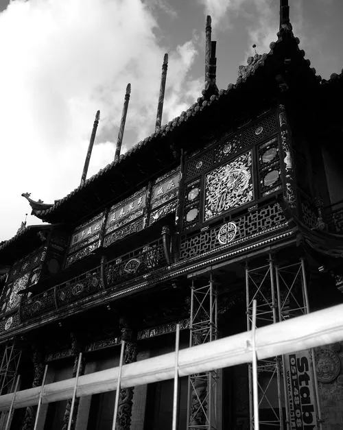 Black and white photo of an ornate traditional Asian building facade with decorative carvings and vertical poles extending from the roof, partially obscured by scaffolding.