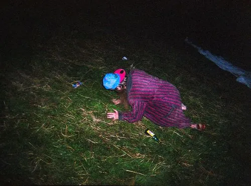 Person in a striped coat and colorful cap lying face down on grass at night next to a bottle.