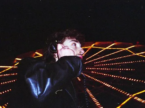 Person in a black leather jacket holding a telephone receiver with a Ferris wheel lit up at night in the background.