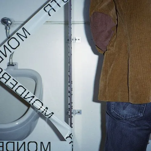 Person wearing a brown corduroy jacket and blue jeans standing next to a urinal in a restroom with a tiled wall.