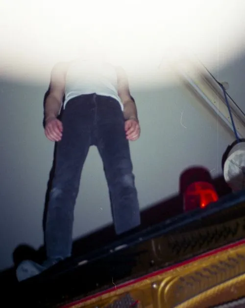 Low-angle view of a person in a white tank top and dark pants standing with hands by their sides, partially obscured by bright light above.