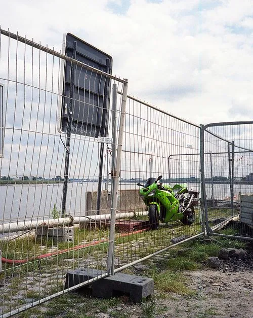 Bright green motorcycle parked behind a wire fence near a river with a cloudy sky.