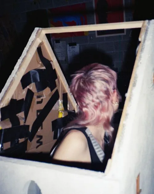 Person with short pink hair looking away, sitting inside a cardboard structure partially covered with black tape.