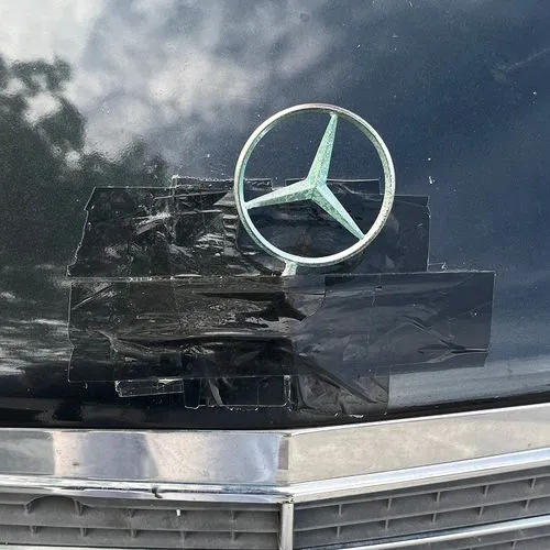 Close-up of a black car hood with a Mercedes-Benz emblem held on by multiple pieces of black tape above the front grille.