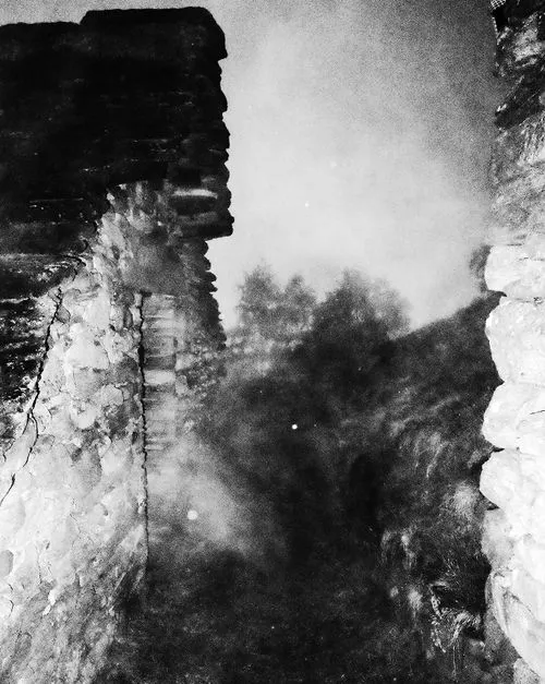 Black and white image of old, rough stone walls with mist or smoke creating an eerie atmosphere and blurry trees in the background.
