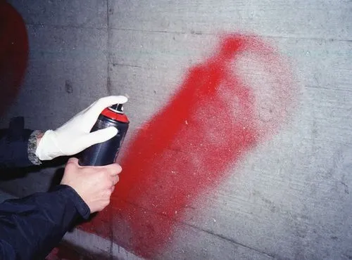 Person wearing white gloves spraying red paint from a can onto a gray concrete wall.