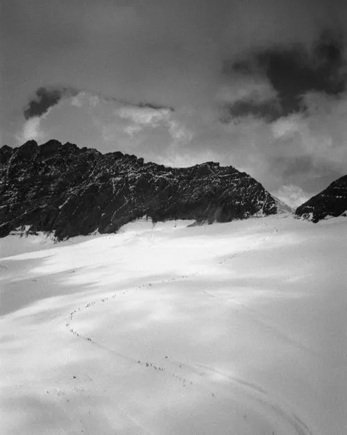 Black and white photo of a large group of climbers trekking in a line across a snowy mountain slope beneath rugged rock formations.