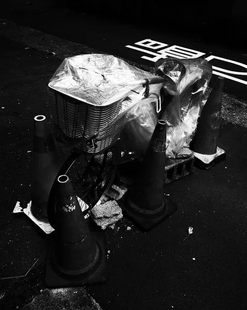 Black and white photo of a bicycle covered in plastic sheets secured with tape, surrounded by four traffic cones on the pavement.