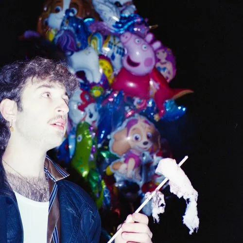 Man with curly hair and mustache holding partially eaten pink cotton candy, colorful character balloons in the dark background.