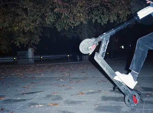 Close-up of a person performing a wheelie on an electric scooter on a leaf-strewn pavement at night.