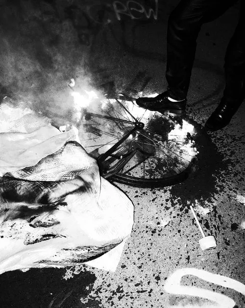 Black-and-white photo showing a burning bicycle wheel on the ground with a person’s legs and polished shoes nearby.