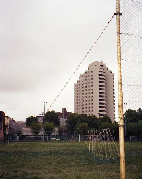 Soccer field with goalpost and net in the foreground, large modern apartment building and trees in the background under an overcast sky.