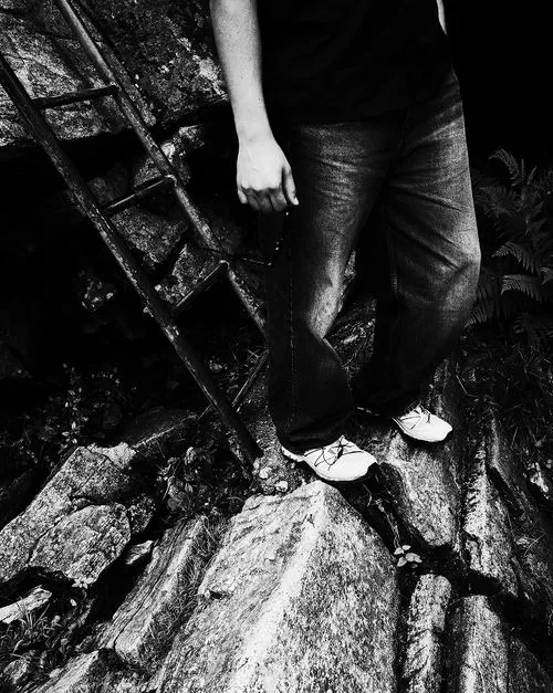 Close-up of a person standing on uneven rocks next to a rusty ladder, wearing jeans and white shoes.