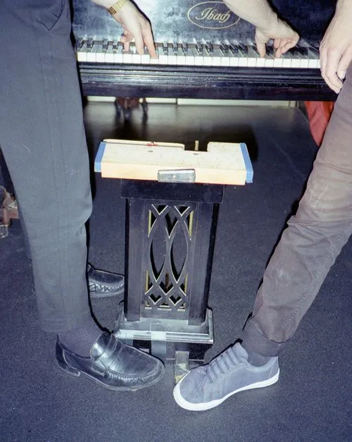 Two people playing an Ibach piano with their hands on the keys and their feet near the pedals.