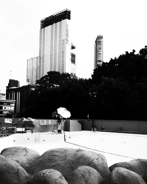 Empty outdoor swimming pool with large rocks in foreground and tall buildings and trees in the background.