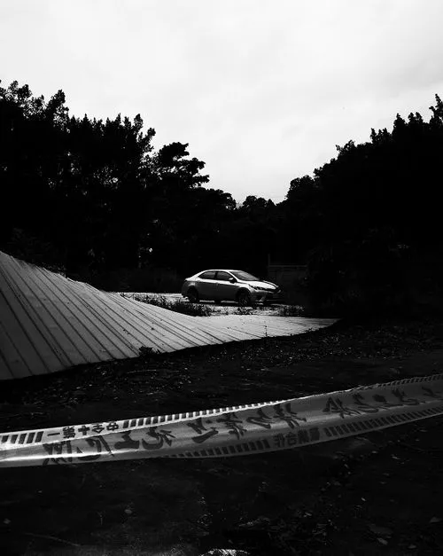 Silver sedan parked near fallen metal roofing sheets, cordoned off with warning tape, with trees in background under a cloudy sky.