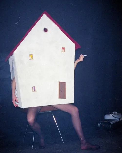 Person sitting on a chair wearing a house costume, with bare legs and arms, holding a cigarette in one hand against a dark background.