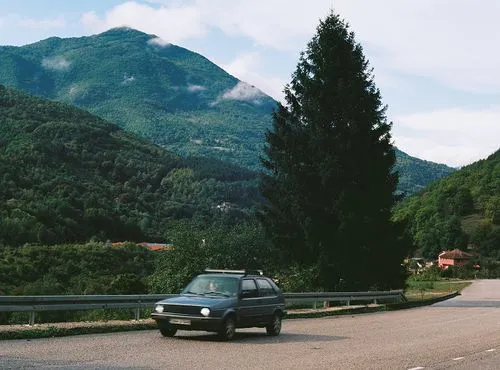 Small car driving on a curving road surrounded by lush green hills and large conifer tree under a partly cloudy sky.