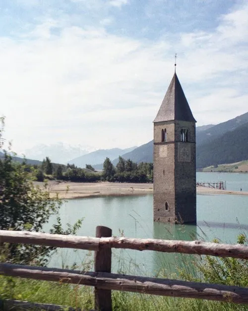 Stone bell tower partially submerged in a lake with mountains and trees in the background under a cloudy sky.